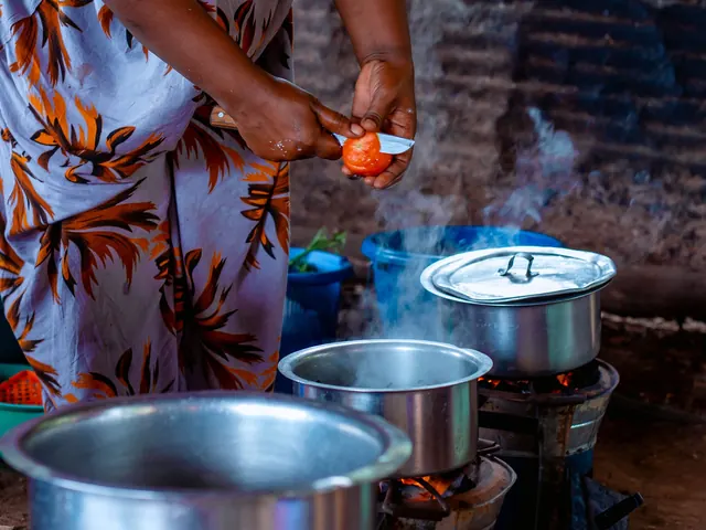 A cook peels tomatoes beside steaming pots on charcoal stoves